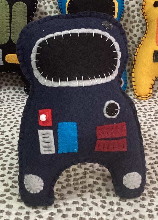 A handmade felt astronaut plush toy in dark navy blue, featuring a large black visor with white stitched details, a red button, a blue panel, and red and white accents, resting on a patterned fabric surface.