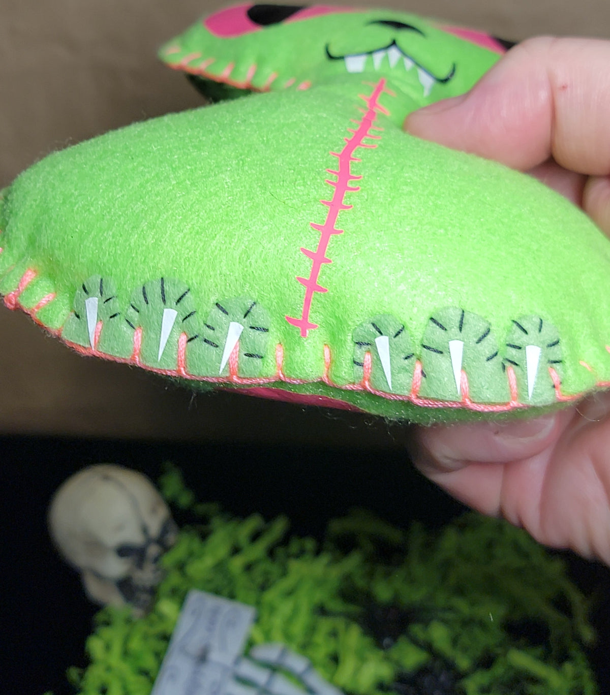 A hand holding a bright green felt monster plush toy with pink stitched details and embroidered teeth, shown against a dark background with decorative props.