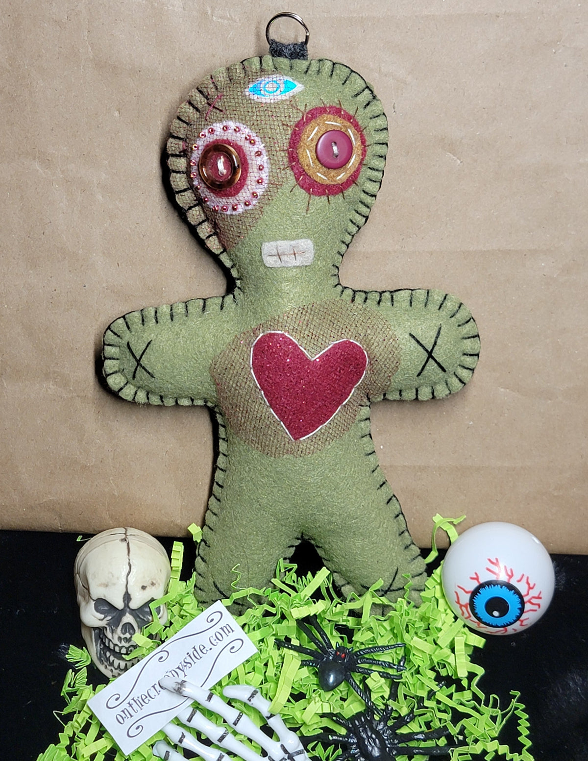 A handmade green fabric voodoo doll ornament with colorful embroidered details, displayed on a black surface with skull, skeleton hand, spider, and eyeball decorations against a brown cardboard background.