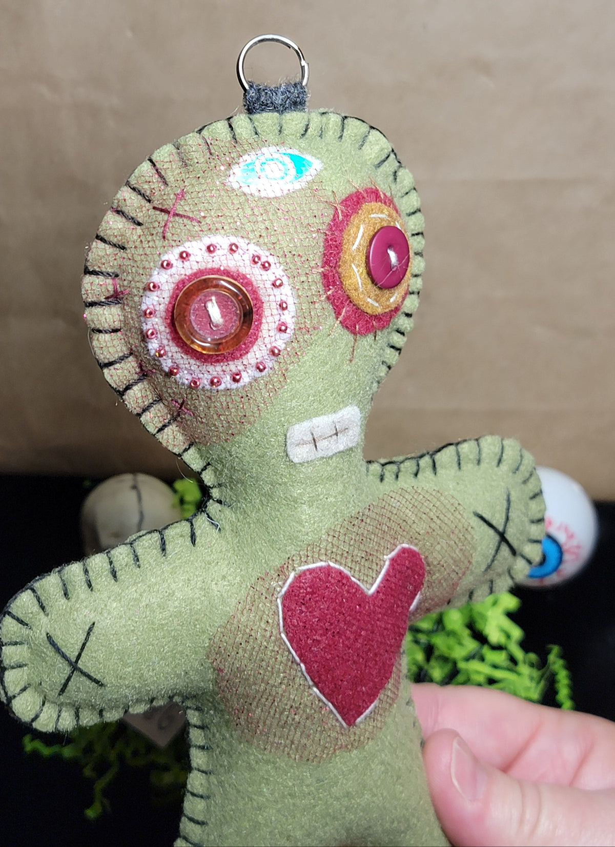 A handmade fabric voodoo doll hanging ornament in sage green, held by a hand against a brown paper background. The doll features large button eyes, a stitched mouth, and a red heart on its chest.