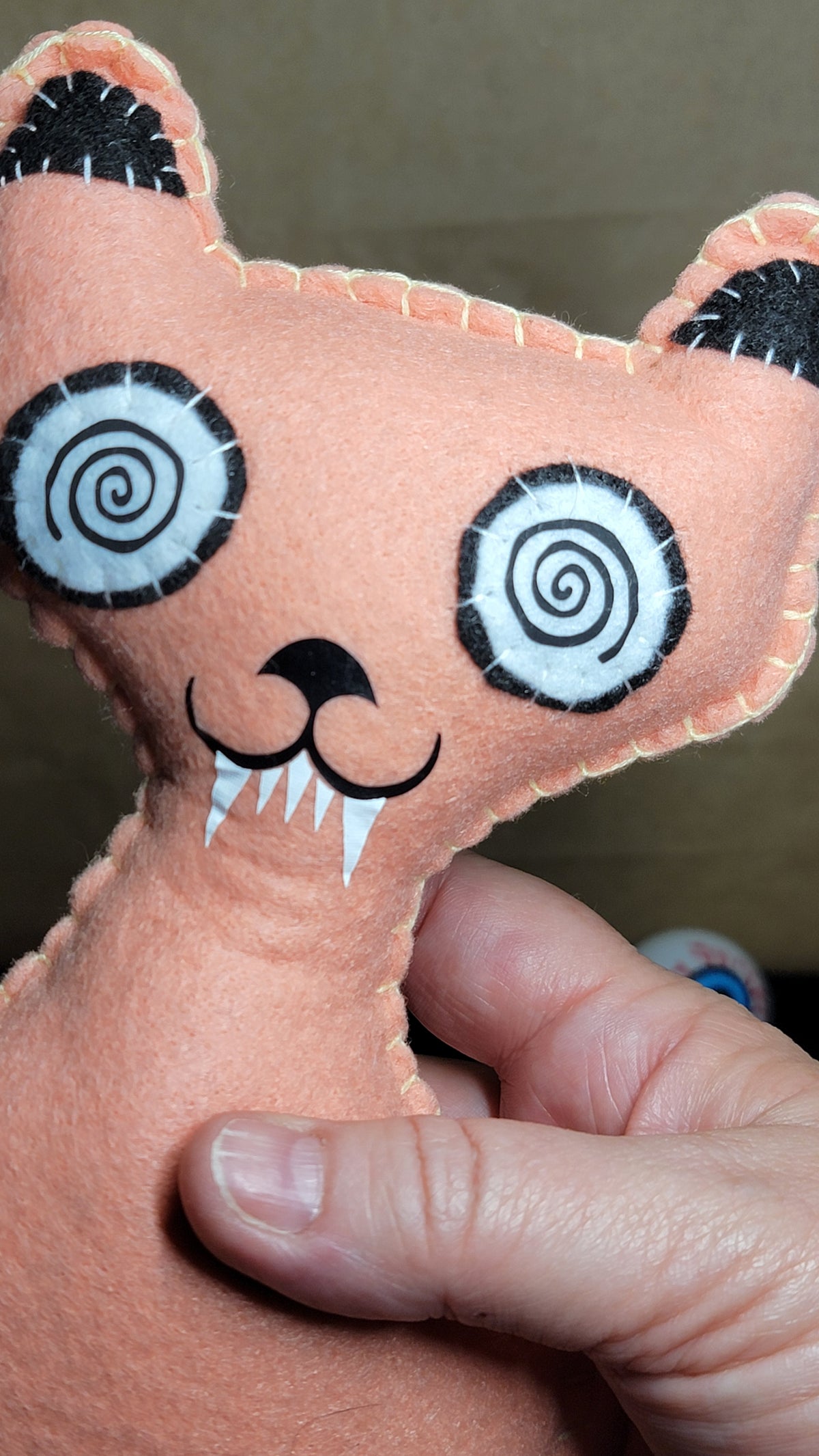 A close-up of a handmade peach-colored plush cat toy being held by a hand. The cat has black and white spiral eyes, a black stitched nose, and white fangs.