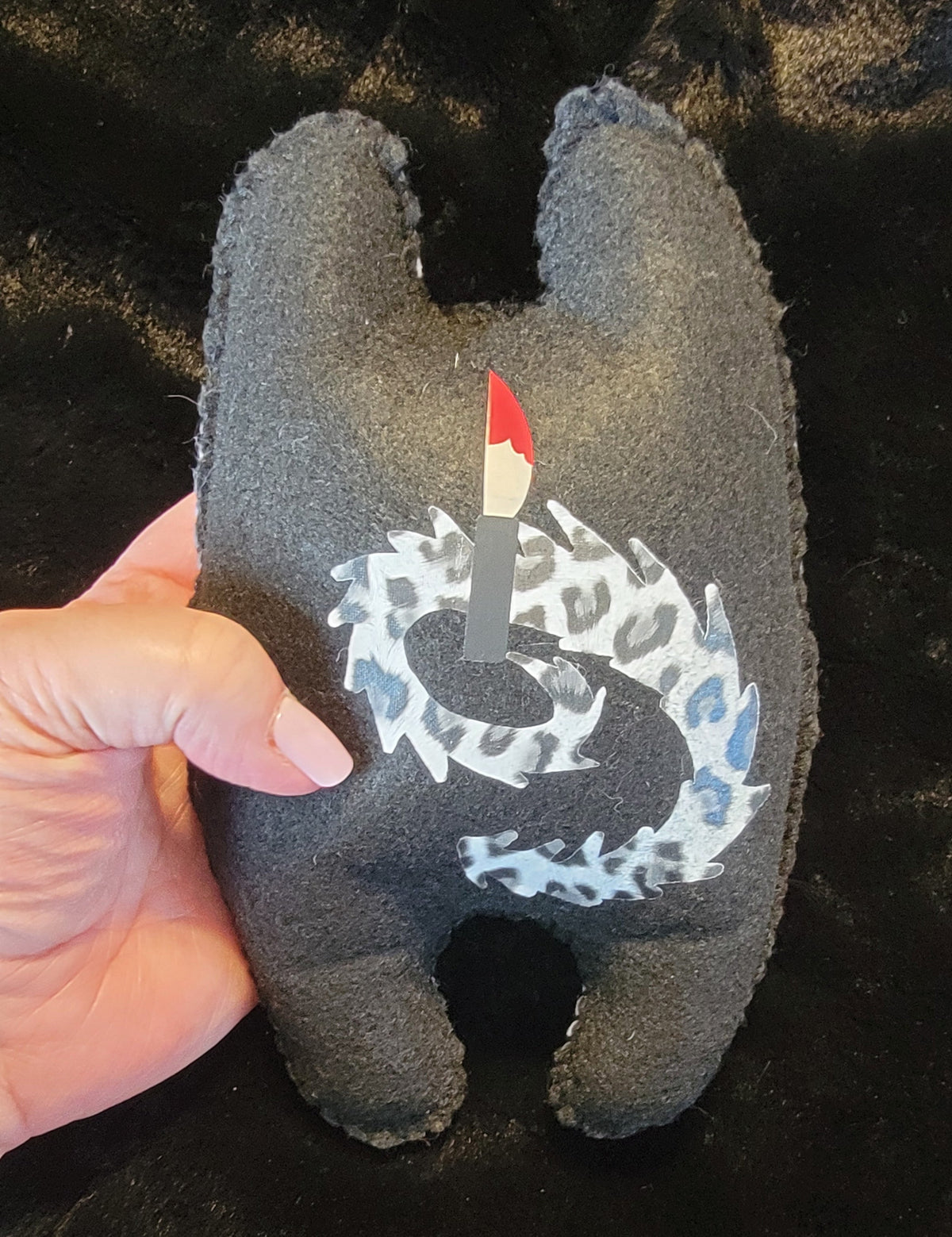 A hand holding a dark gray plush tooth-shaped toy with a white and blue dragon design and a small red-and-black candle on the front, set against a black textured background.