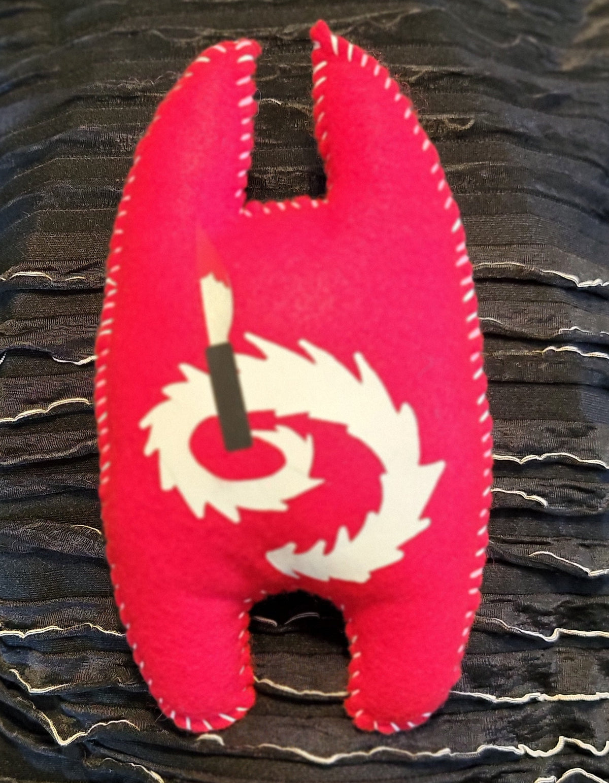 A red felt H-shaped brooch featuring a white spiral design and a black center pin, displayed on a dark textured background.