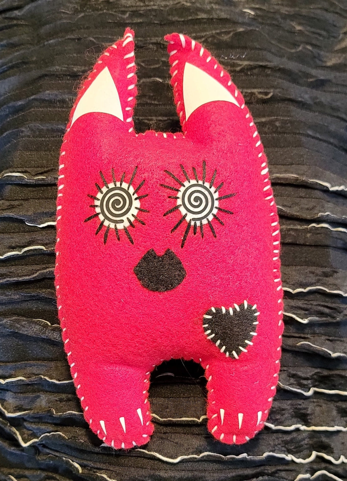 A handmade felt cat-shaped brooch in bright pink with embroidered black and white details, displayed on a black textured pleated fabric background.