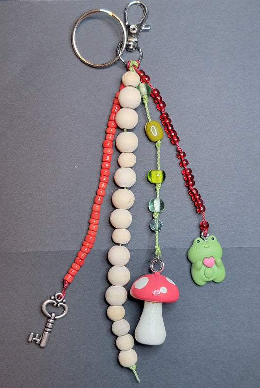 A handmade keychain featuring a silver split ring, a red beaded chain with a silver key charm, a vertical strand of cream-colored wooden beads, a red beaded chain with a green frog charm, and a red and white mushroom charm, all laid out on a gray background.