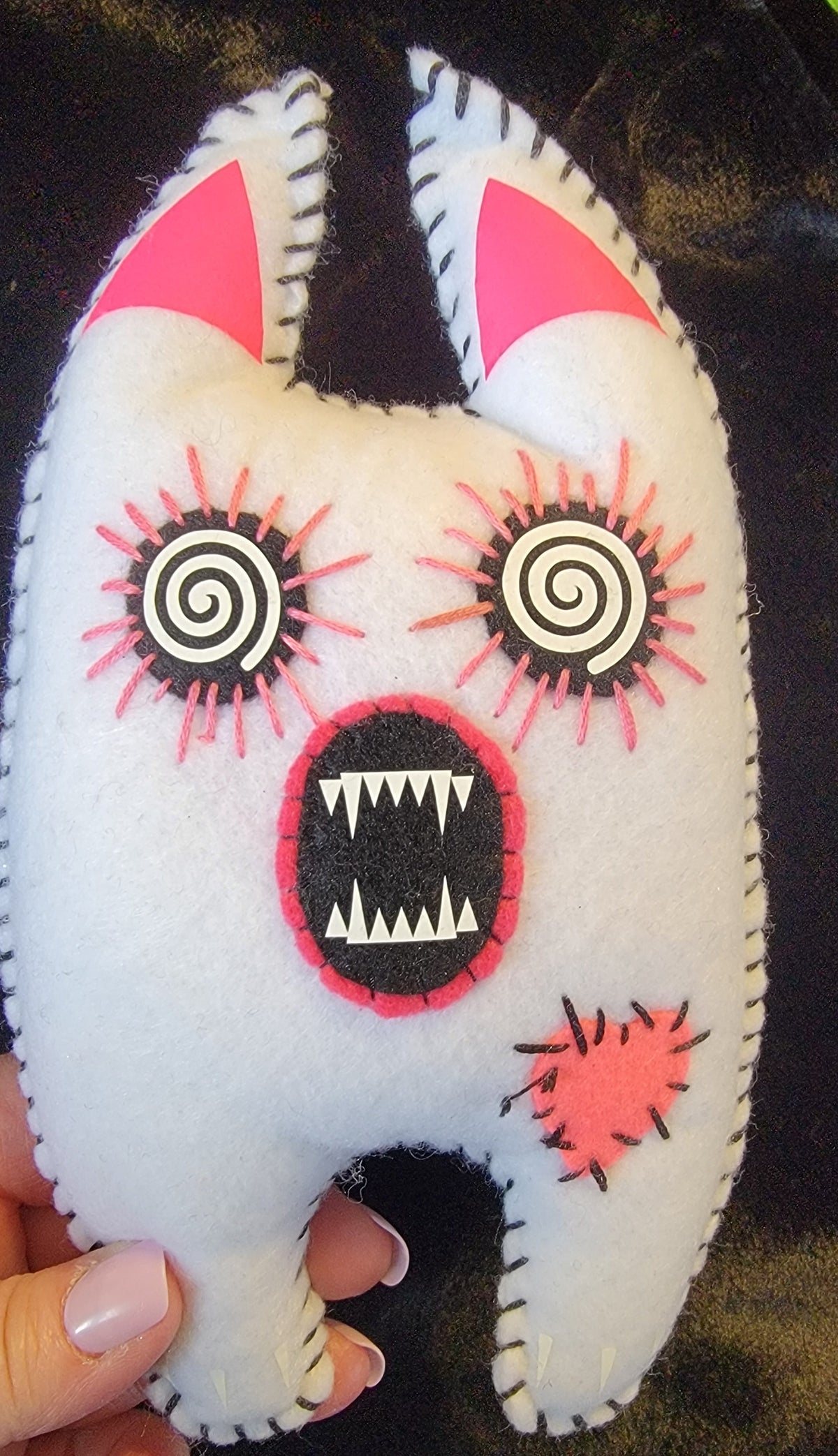 A handmade white felt monster toy with pink and black embroidered details, held by a hand against a dark textured background.