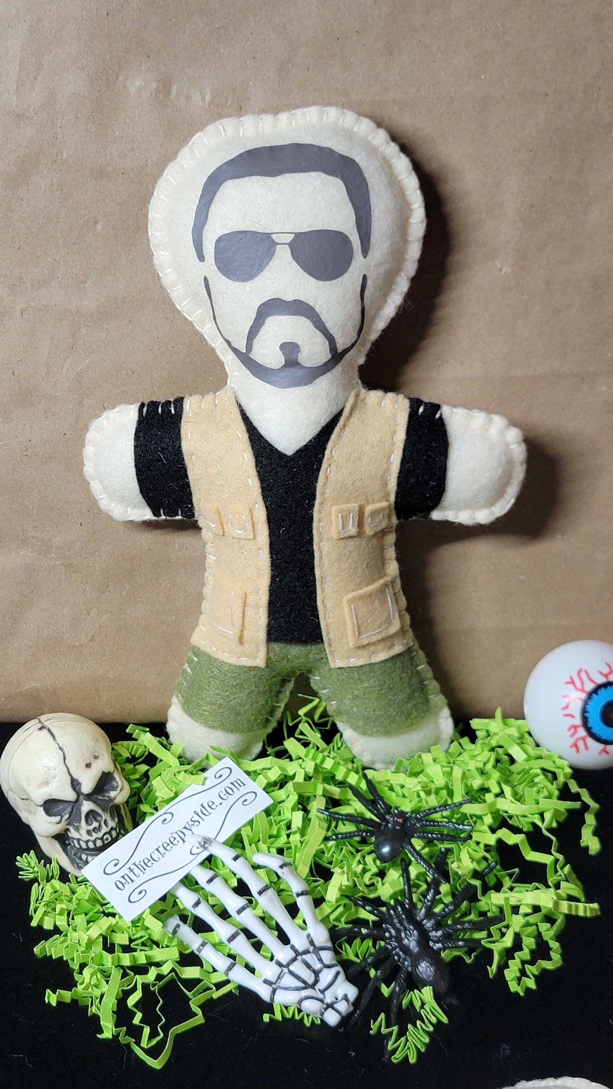 A handmade felt doll of Heisen Wiker, styled with a skull, skeleton hand, plastic spiders, and bright green shredded paper on a brown cardboard background.