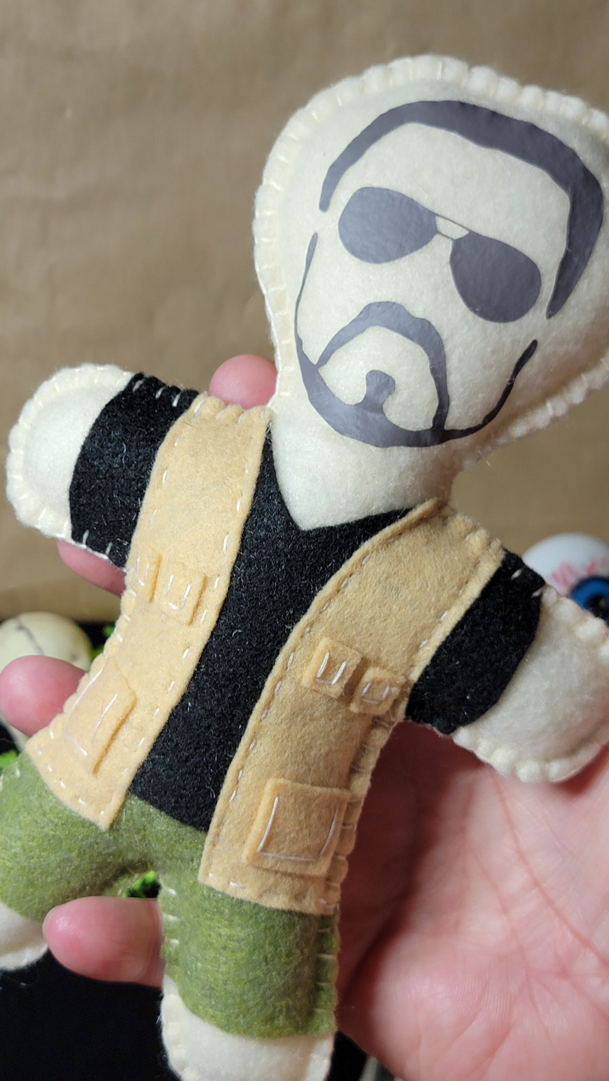 A handmade felt doll being held in a hand, featuring a printed face with sunglasses and a beard, wearing a black shirt, tan vest, and green pants.