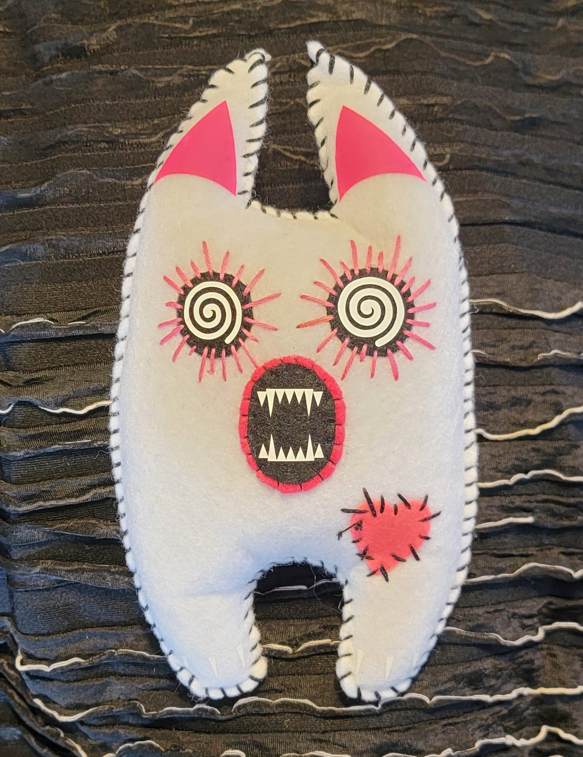 A handmade white felt monster patch with pink ears, spiral eyes, and an open mouth showing teeth, placed on a black ruffled fabric background.