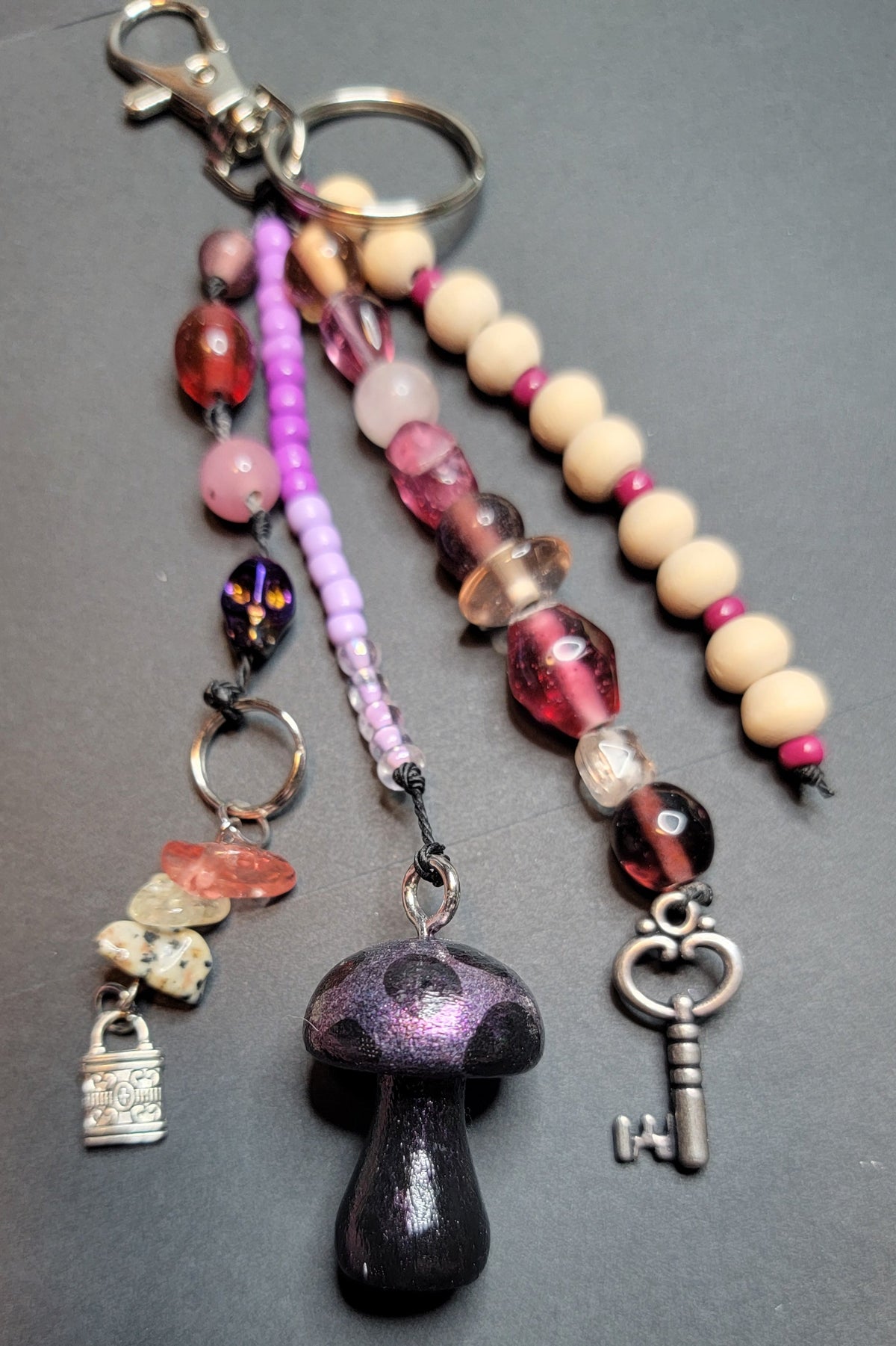A beaded keychain charm set with a large dark purple mushroom pendant, a silver key, a small silver padlock charm, pink and purple beads, wooden beads, and translucent pink gemstones, displayed on a dark gray surface.