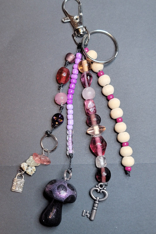A beaded keychain with multiple hanging strands, including pink and purple beads, cream wooden beads, a black mushroom charm, a silver key, and a small lock charm, displayed on a dark gray background.