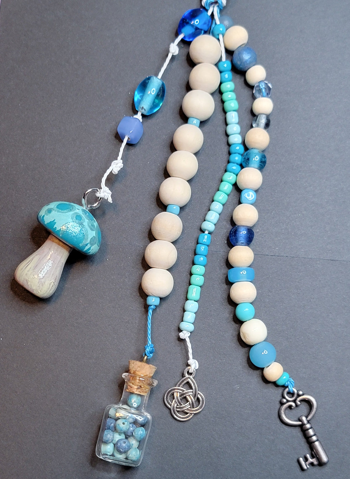 A beaded charm or tassel accessory laid flat on a dark gray background, featuring cream and blue beads, a turquoise-and-white mushroom charm, a small glass bottle filled with blue beads, a silver knot charm, and a silver key.
