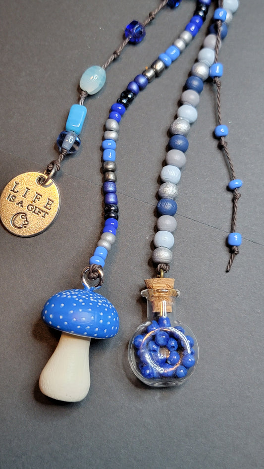 Three beaded cords with blue and white beads, displayed on a dark gray background. One cord has a mushroom-shaped pendant, one has a small glass vial containing blue beads, and one has a round gold-toned charm engraved with 'LIFE IS GIFT'.