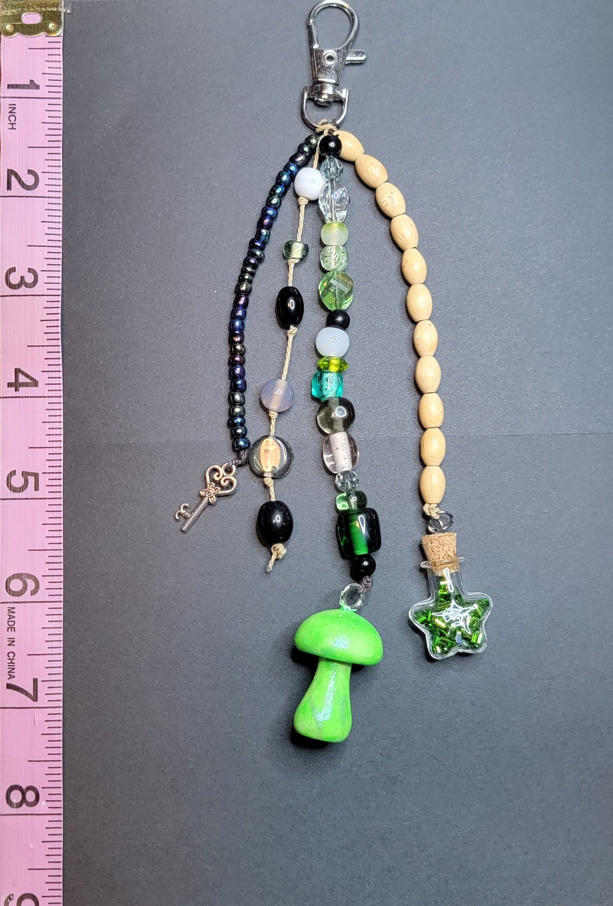 A handmade beaded keychain or bag charm with multiple strands, including a bright green mushroom pendant, a green star charm, cream-colored beads, black beads, and a silver clasp, shown next to a pink measuring ruler on a dark gray background.