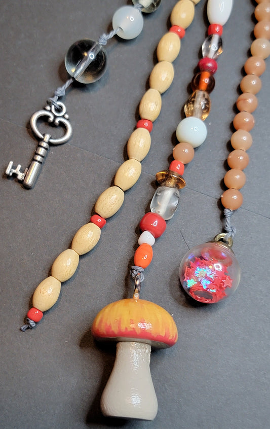 A close-up of two beaded necklaces laid on a dark gray surface. One necklace features light wood beads with small red spacer beads and a silver key charm.