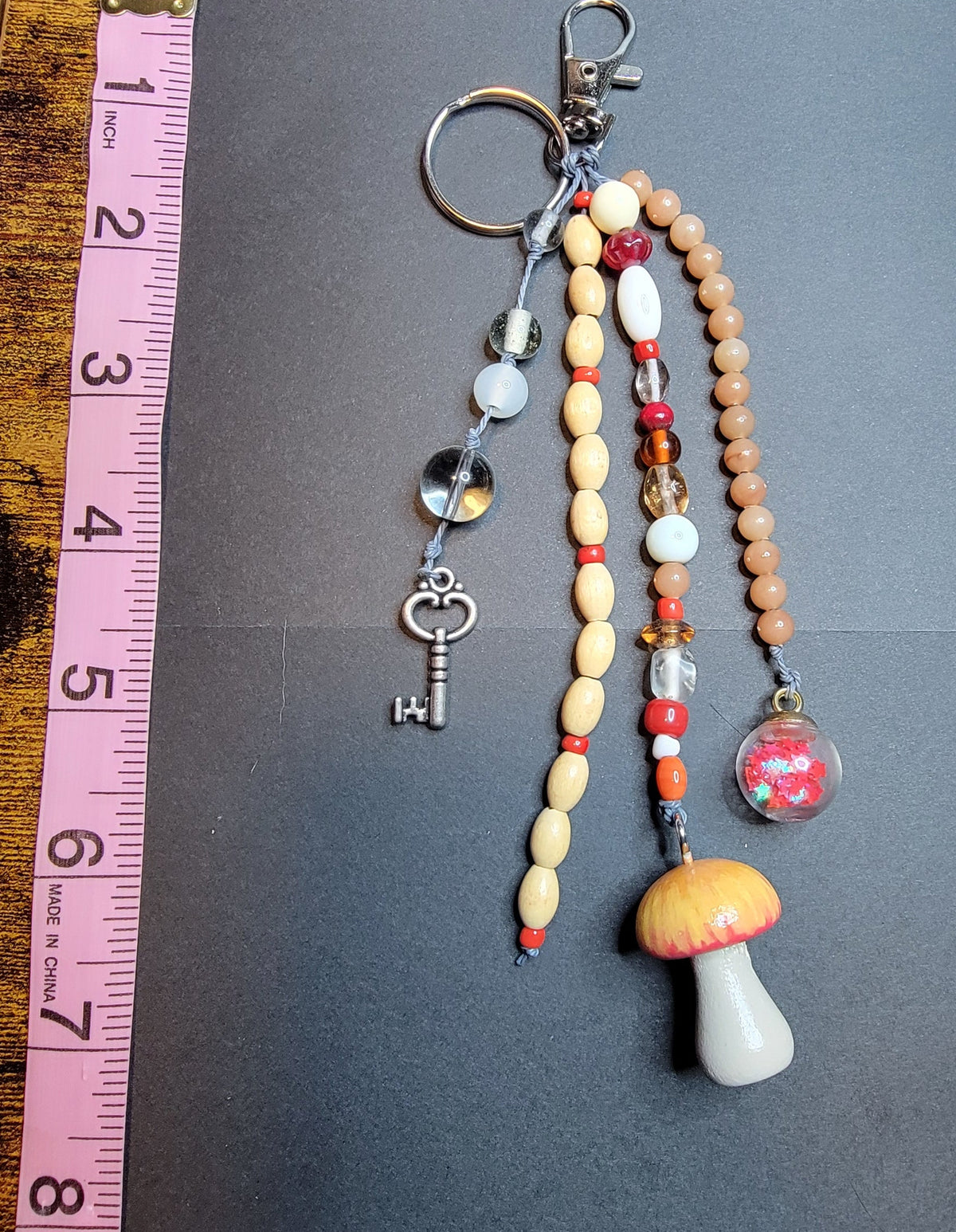 A handmade beaded keychain with a mushroom charm, key, and glass beads, displayed on a dark gray card next to a pink measuring tape on a wooden table.