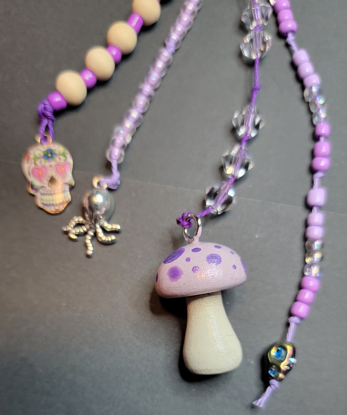 A close-up of a beaded necklace with a mushroom pendant, purple beads, clear faceted beads, a silver octopus charm, and a skull charm, laid on a dark gray surface.