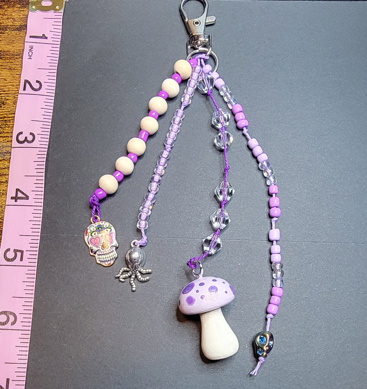 A purple and white beaded phone strap with a mushroom charm and silver clasp, laid on a black card over a wooden table with a pink measuring tape visible.
