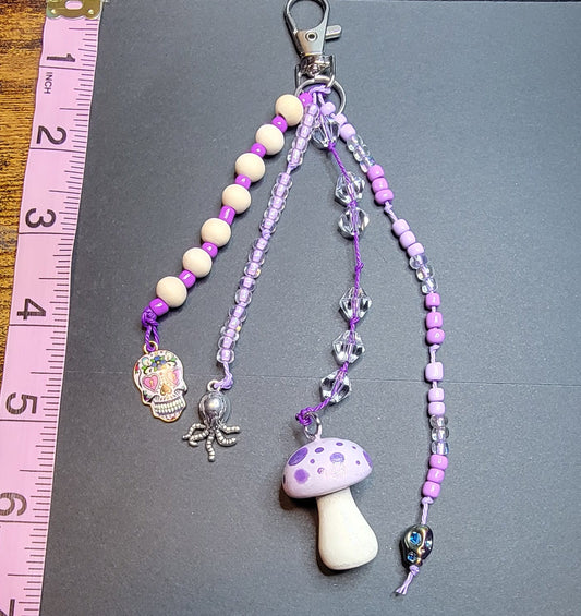 A purple and white beaded phone strap with a mushroom charm and silver clasp, laid on a black card over a wooden table with a pink measuring tape visible.