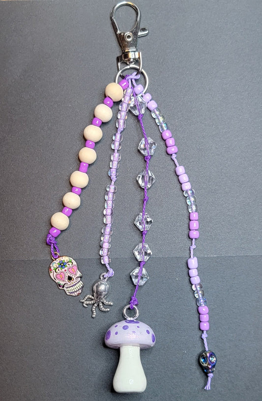 A purple and lavender beaded keychain or phone strap with a silver lobster clasp, featuring a mushroom charm, a sugar skull charm, a small octopus charm, and clear faceted beads on a dark gray background.
