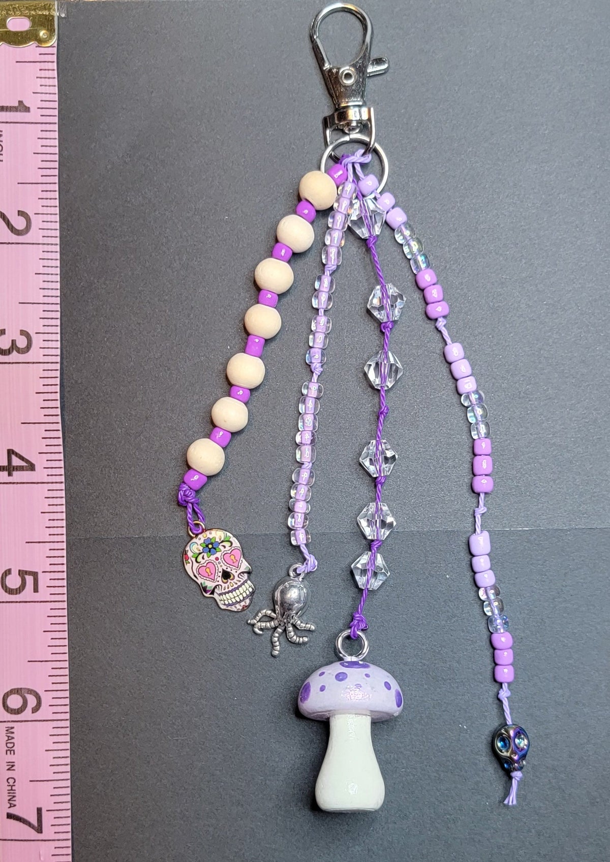 A handmade beaded keychain or bag charm with a silver lobster clasp, featuring purple and lavender beads, a small mushroom charm, and a pink spider charm, displayed on a dark gray card with a pink measuring tape on the left side.