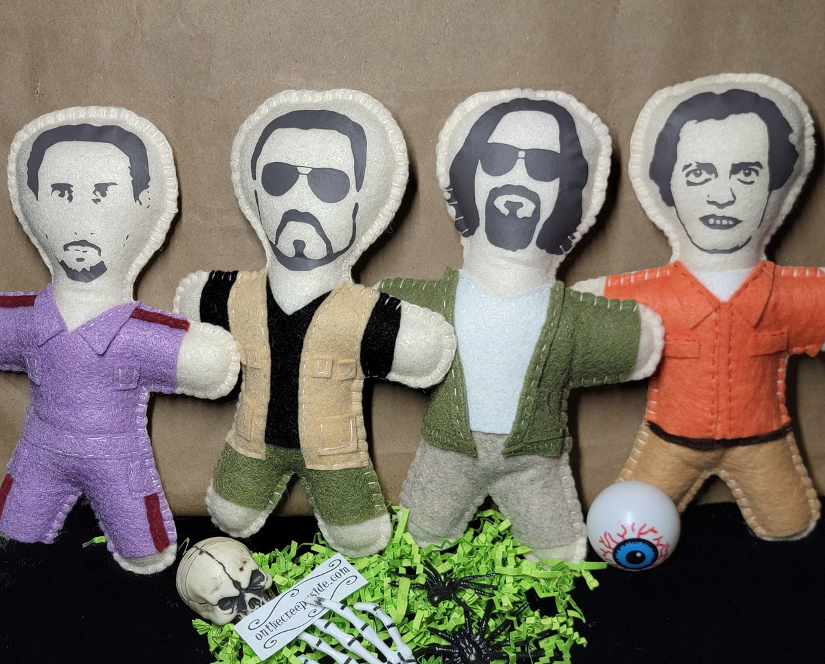 Four fabric doll figures with printed faces and body garments in purple, black-and-beige, green-and-white, and orange, arranged on a brown backdrop with a black surface below. A small skull, green shredded paper, and a tag are placed in front.