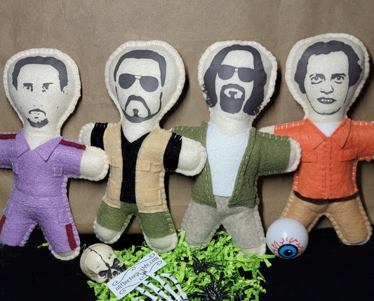 Four fabric doll figures with printed faces and body garments in purple, black-and-beige, green-and-white, and orange, arranged on a brown backdrop with a black surface below. A small skull, green shredded paper, and a tag are placed in front.