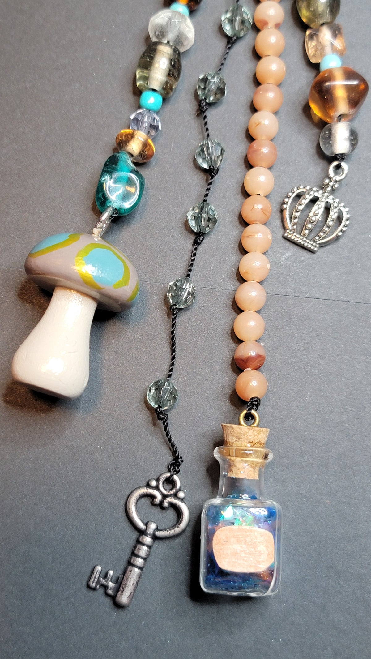 A close-up of a beaded necklace laid flat on a dark gray surface. The necklace features peach and cream beads, a turquoise and green mushroom charm, a silver crown pendant, a silver key charm, and a small glass bottle charm filled with blue and gold elements.