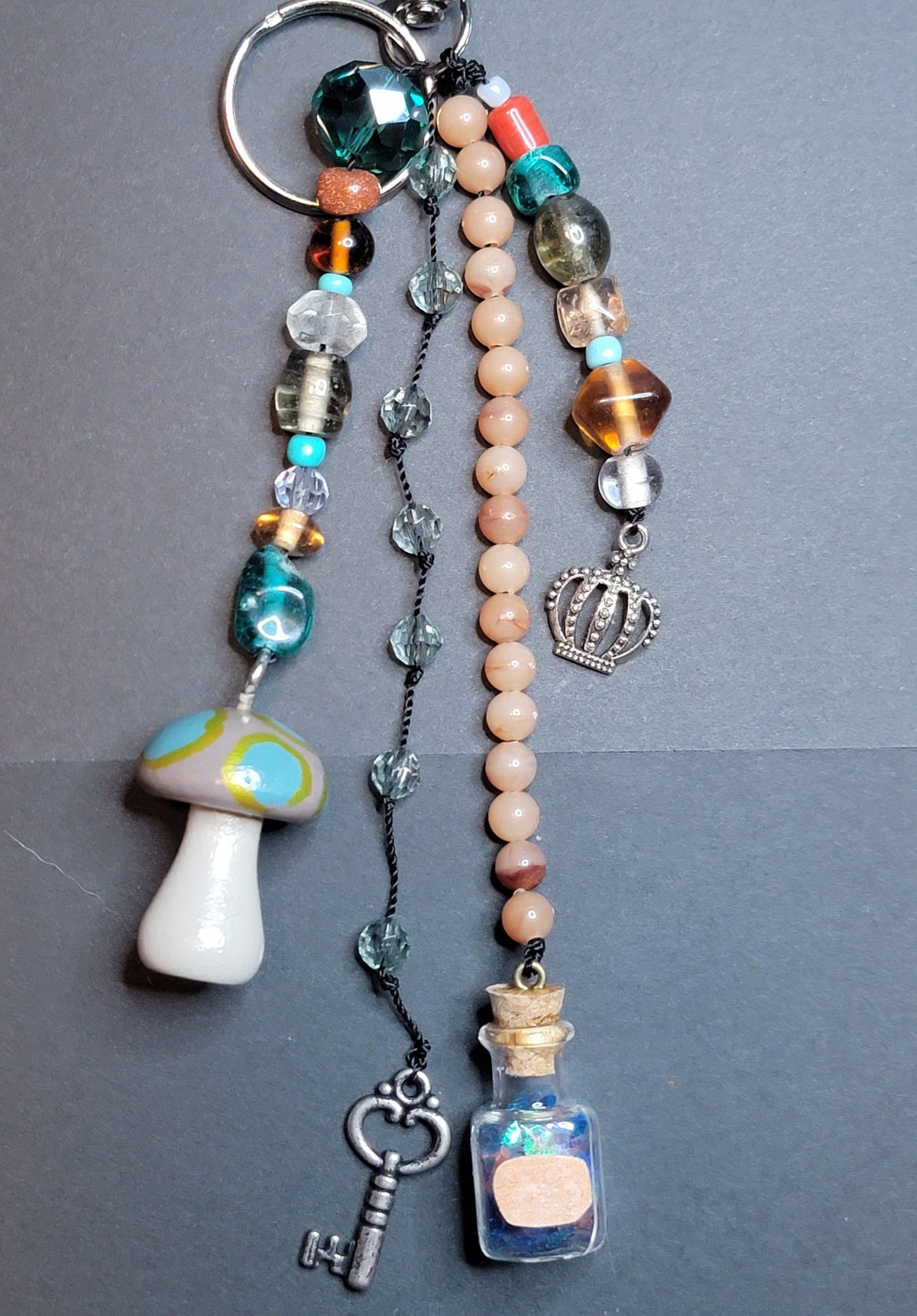 A beaded keychain charm set with a silver split ring, featuring a white mushroom bead, a pink beaded strand, a small glass bottle charm filled with blue and pink sand, a silver key, and assorted colorful beads on a dark gray background.