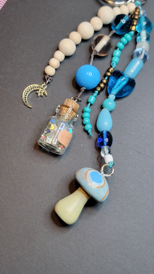 A close-up of a beaded necklace laid on a dark gray surface. The necklace features cream-colored wooden beads, turquoise and blue glass beads, a small glass bottle charm filled with glitter, a crescent moon charm, and a large light blue and cream mushroom-shaped bead.