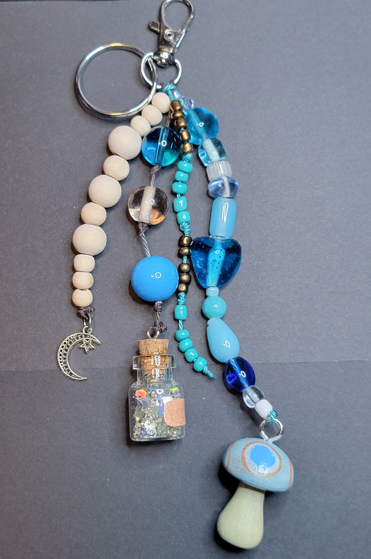 A handmade beaded keychain charm set on a dark gray background. The keychain features a silver split ring, light wood beads, a silver crescent moon charm, a small glass bottle filled with glitter, a light blue heart-shaped bead, turquoise beads, a blue glass bottle, a mushroom-shaped bead, and a blue glass teardrop bead.