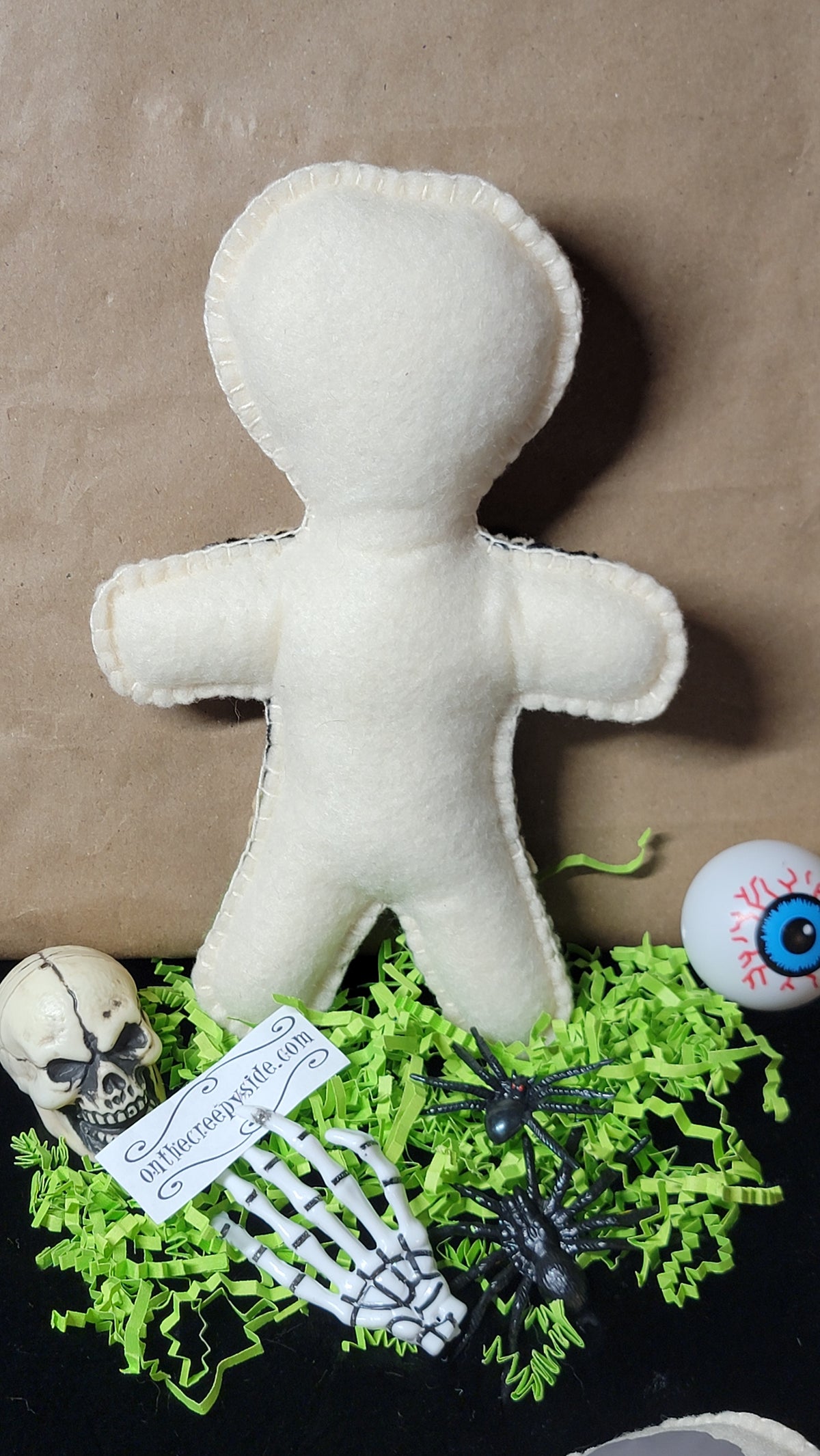 A white felt voodoo doll with a blank face, posed upright on a black surface with green shredded paper, a small skull, a skeleton hand, a spider, and a eyeball prop around it against a brown paper background.