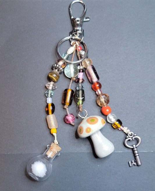 A whimsical mushroom-themed keychain with a silver lobster clasp, featuring a white mushroom charm, a small glass bottle with white powder and cork, colorful beads, and a vintage key on a dark gray background.