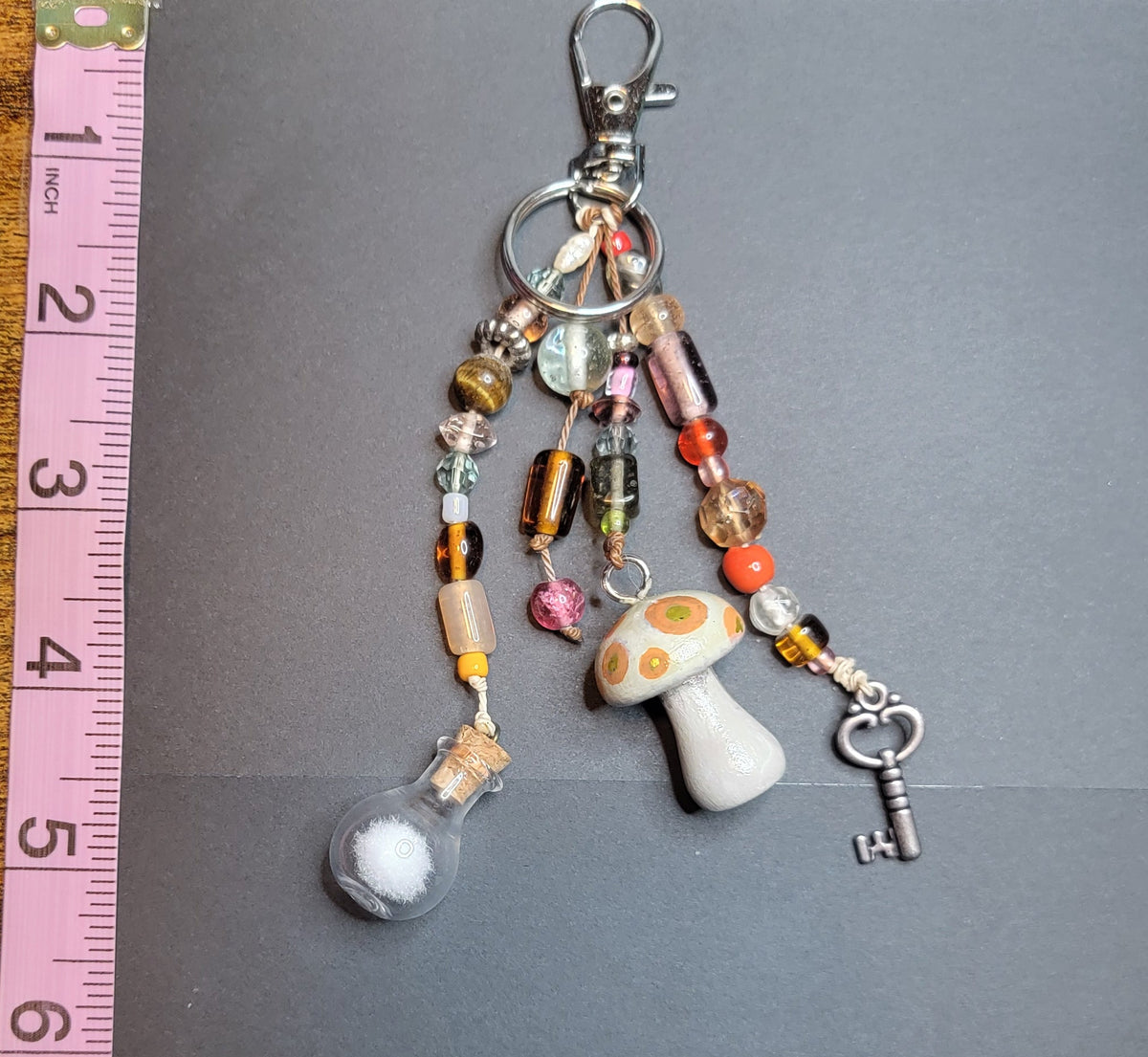 A handmade beaded keychain charm with a silver lobster clasp, featuring colorful glass beads, a small white mushroom charm, a clear bubble bubble, and a silver key, displayed next to a pink measuring tape on a dark gray background.